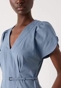 Lichtblauwe jurk met een diepe V-hals, korte puffmouwen en een ceintuur in de taille. Gemaakt van een gladde stof, met subtiele textuur en stikseldetails.