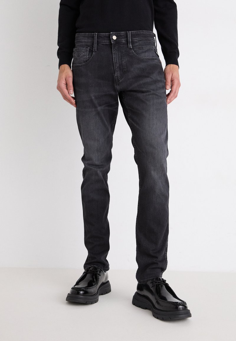 Fakó fekete farmernadrág slim fit szabással, öt zsebével, varrott részletekkel és fémből készült gombos záródással. Fényes fekete cipővel viselve.