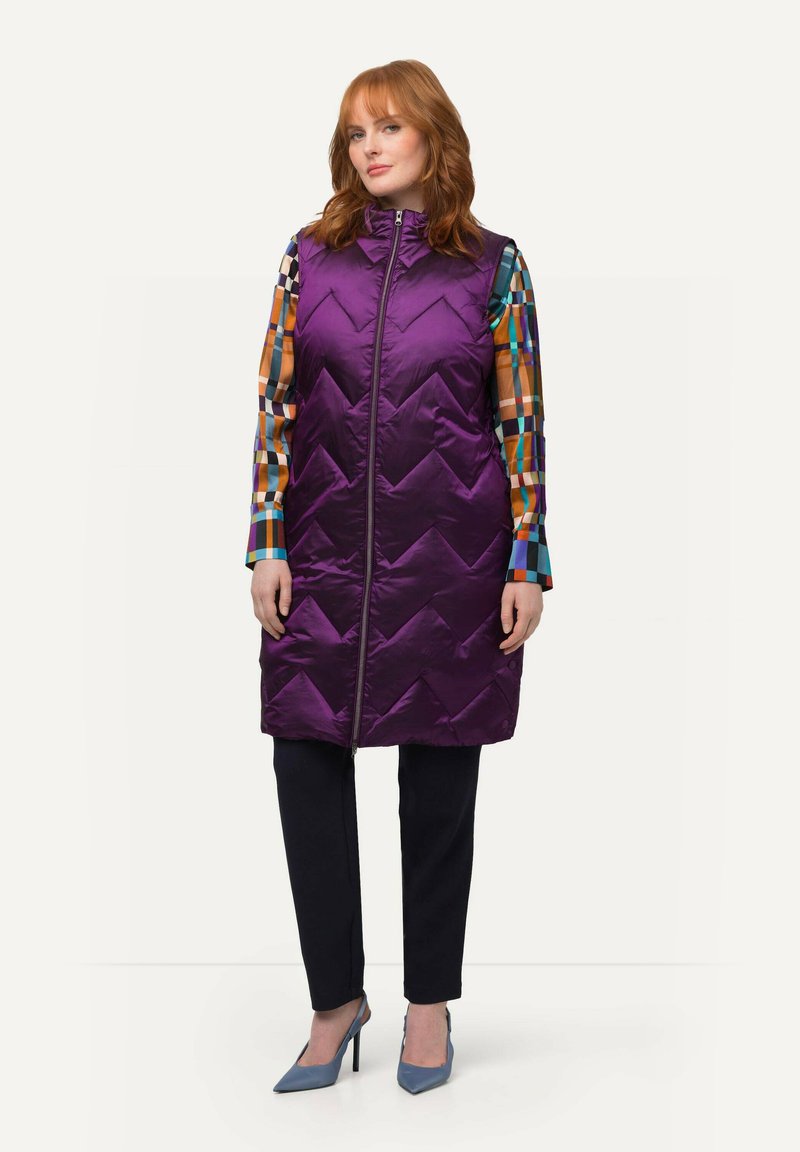 Fialový prošívaný dlouhý vest s vysokým límcem a předním zipem, nosí se přes barevnou kostkovanou dlouhou košili s dlouhým rukávem a černé kalhoty.