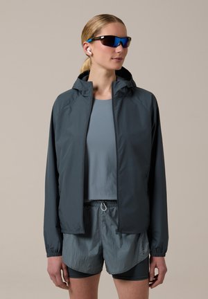 Mujer con chaqueta oscura con cremallera, camiseta deportiva azul, pantalones cortos de varias capas, gafas de sol y auriculares inalámbricos sobre un fondo liso.