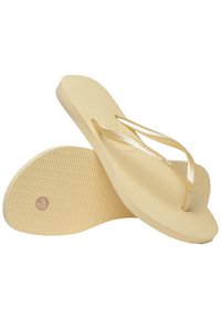 Gule flip-flops med teksturerte såler og glinsende remmer. Sålene har et ribbet mønster og et sirkulært hull nær hælen.