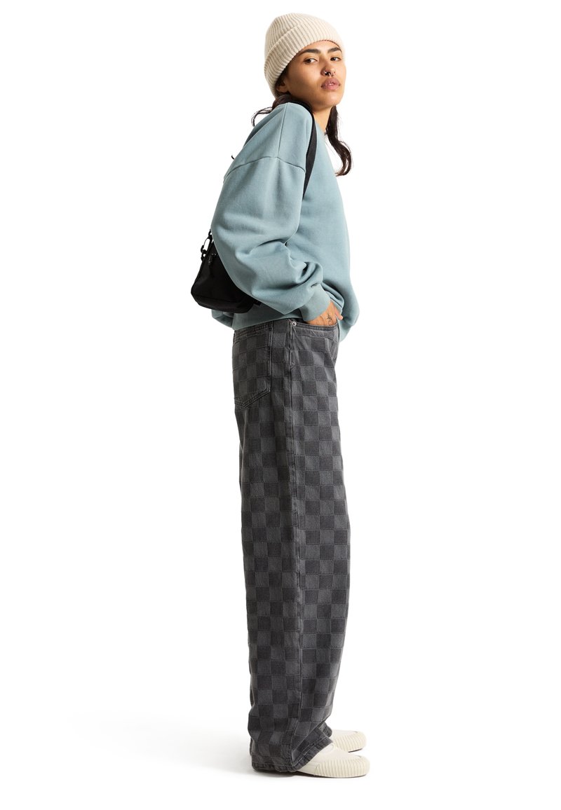 Übergroßer blauer Pullover mit überschnittenen Schultern, dunkle karierte weite Hosen und helle Slipper. Eine schlichte schwarze Tasche rundet das Outfit ab.