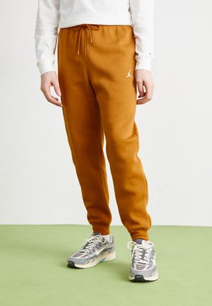 Osoba ubrana w musztardowe spodnie dresowe z białym logo Jordan, białą koszulę z długim rękawem oraz szare buty Nike na zielonej podłodze.