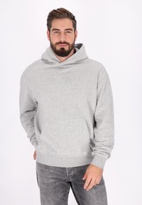 Skäggig man som bär en ljusgrå hoodie och mörkgrå jeans, står med ena handen i fickan mot en ren vit bakgrund.
