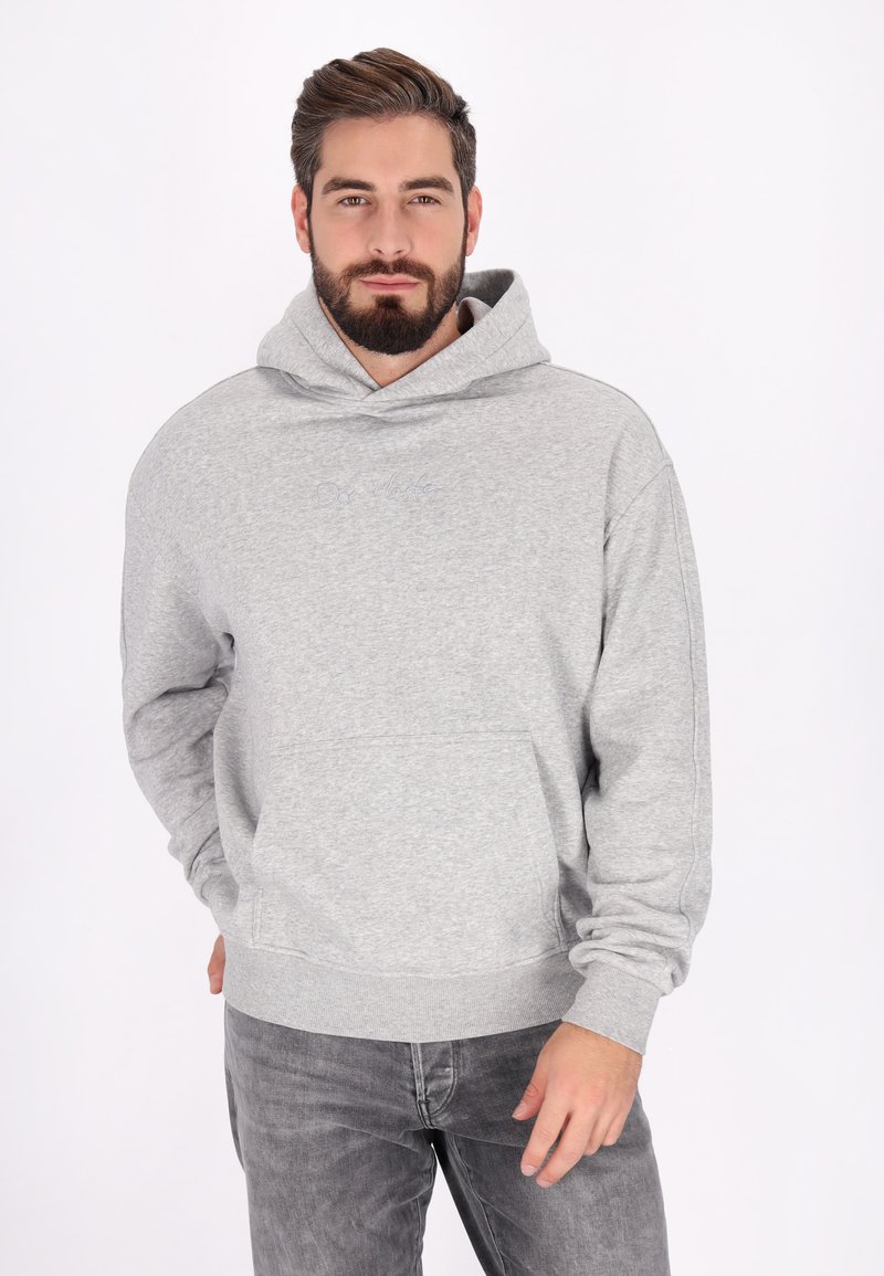 Skäggig man som bär en ljusgrå hoodie och mörkgrå jeans, står med ena handen i fickan mot en ren vit bakgrund.
