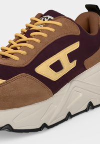 Χοντρό sneaker με μωβ ύφασμα, καφέ σουέντ επικαλύψεις, κίτρινα κορδόνια, κίτρινο λογότυπο στο πλάι και παχύ λευκό πάτο με μαύρες λεπτομέρειες πέλματος.