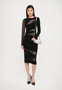 Vestito nero aderente con maniche lunghe trasparenti e pannelli a taglio. Abbinato a una borsa a tracolla argento e tacchi a punta neri.