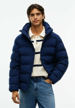 Ung mand iført en navyblå dunjakke over en hvid striktrøje med navyblå striber og blå jeans, stående med den ene hånd i lommen.