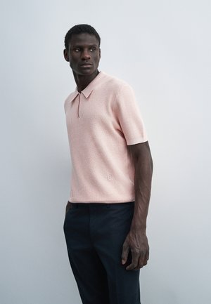 Άνδρας που φοράει ροζ ανοιχτό κοντομάνικο μπλουζάκι πόλο με υφή και σκούρο παντελόνι, στέκεται και κοιτάζει προς τα αριστερά μπροστά σε απλό φόντο.