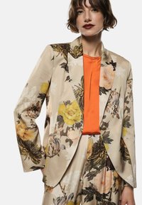 Blazer a fantasia floreale in beige con fiori gialli e crema. Abbinato a una maglietta arancione. Presenta un colletto a revers e chiusura a doppio bottone.