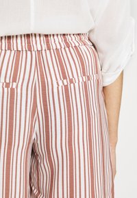 Páskované kalhoty volného střihu v odstínu rezavé a bílé. Mají elastický pas a boční kapsy. Vyrobeny z lehké tkaniny.