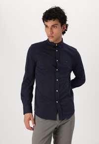 Marineblauwe blouse met lange mouwen van gladde stof, voorzien van een klassieke kraag en witte knopen. Subtiele logo op de borst.