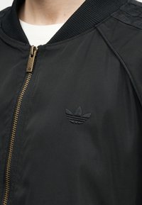 Κοντινό πλάνο μαύρου μπουφάν μπόμπερ με ριμπ κολάρο, χρυσό φερμουάρ και τονικό λογότυπο Adidas trefoil στο στήθος.