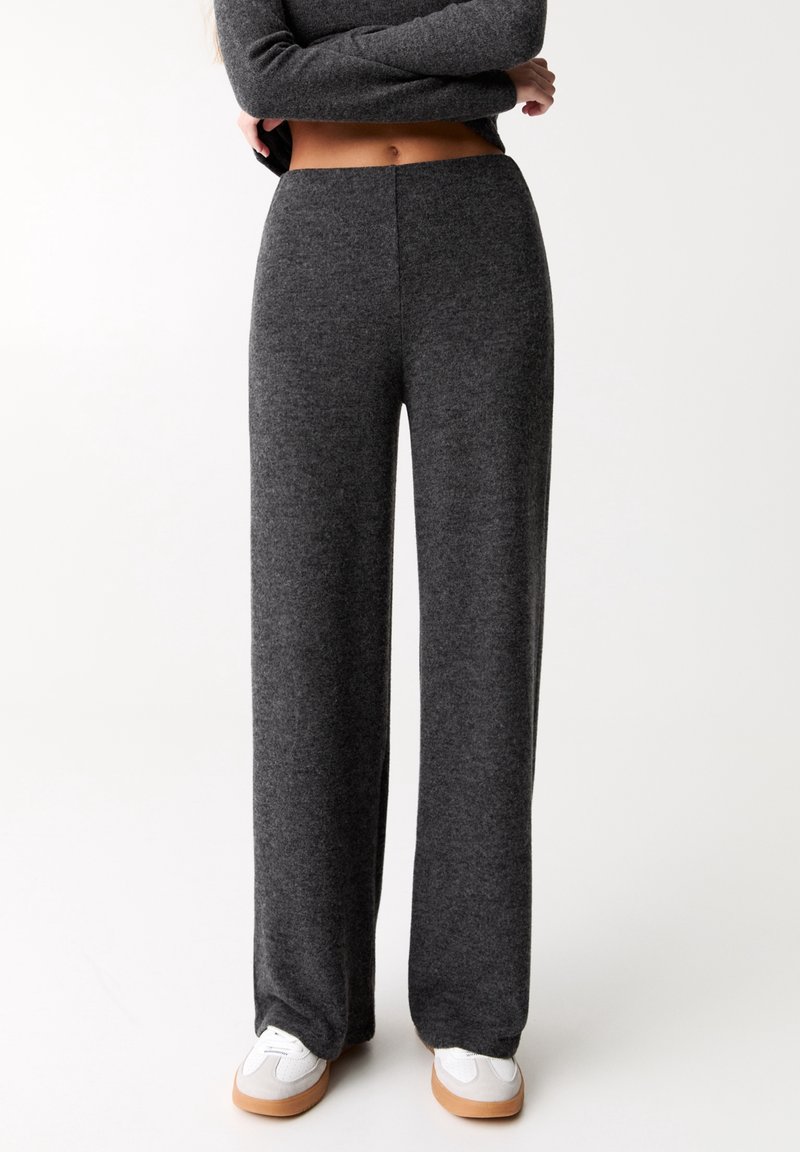 Personne portant un pantalon large gris foncé et des baskets blanches, debout les bras croisés sur le ventre nu.