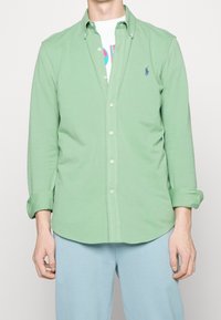 Mężczyzna w jasnobeżowej koszuli z guzikami, z podwiniętymi rękawami, na białym T-shircie z grafiką i jasnoniebieskich spodniach, stojący na tle jednolitego tła.