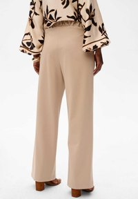 Beige wijduitlopende broek met een gladde textuur, voorzien van een elastische tailleband. Model draagt open schoenen met een houten blokhak.