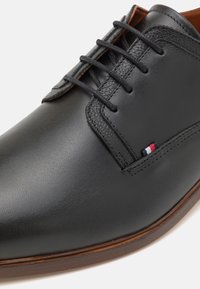 Chaussure habillée noire en cuir avec une texture lisse, accents marron, lacets et un petit détail de logo tricolore. Design de bout concave.