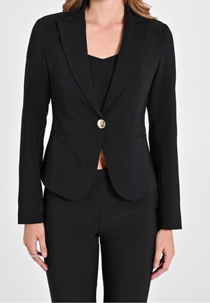 Kvinde iført en tætsiddende sort blazer med en enkelt guldknap, kombineret med sorte bukser og en sort top under.