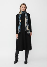 Sort langærmet kjole med en flydende design. Et mønstret tørklæde med blå og beige detaljer svøber sig omkring halsen. Tætsiddende sorte støvler.