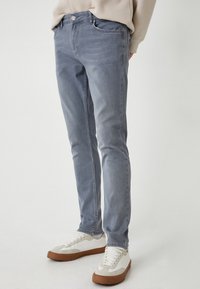 Jeans de denim gris claro con un corte ajustado, que cuentan con un diseño de cinco bolsillos, cierre frontal con botón y detalles de textura sutiles.