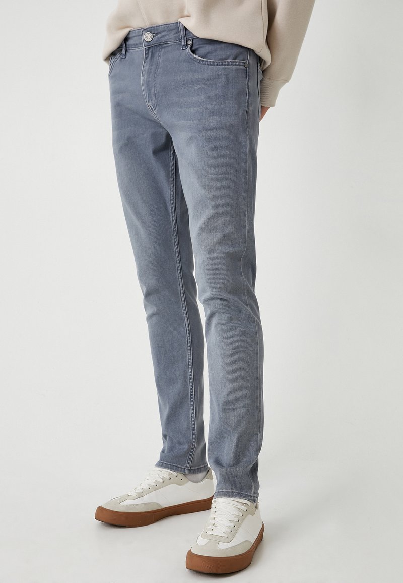 Jeans de denim gris claro con un corte ajustado, que cuentan con un diseño de cinco bolsillos, cierre frontal con botón y detalles de textura sutiles.