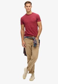 Camiseta de cuello redondo texturizada en rojo, chinos color crema y zapatillas beige. Una camisa de cuadros está atada a la cintura. Conjunto casual con un diseño sencillo.