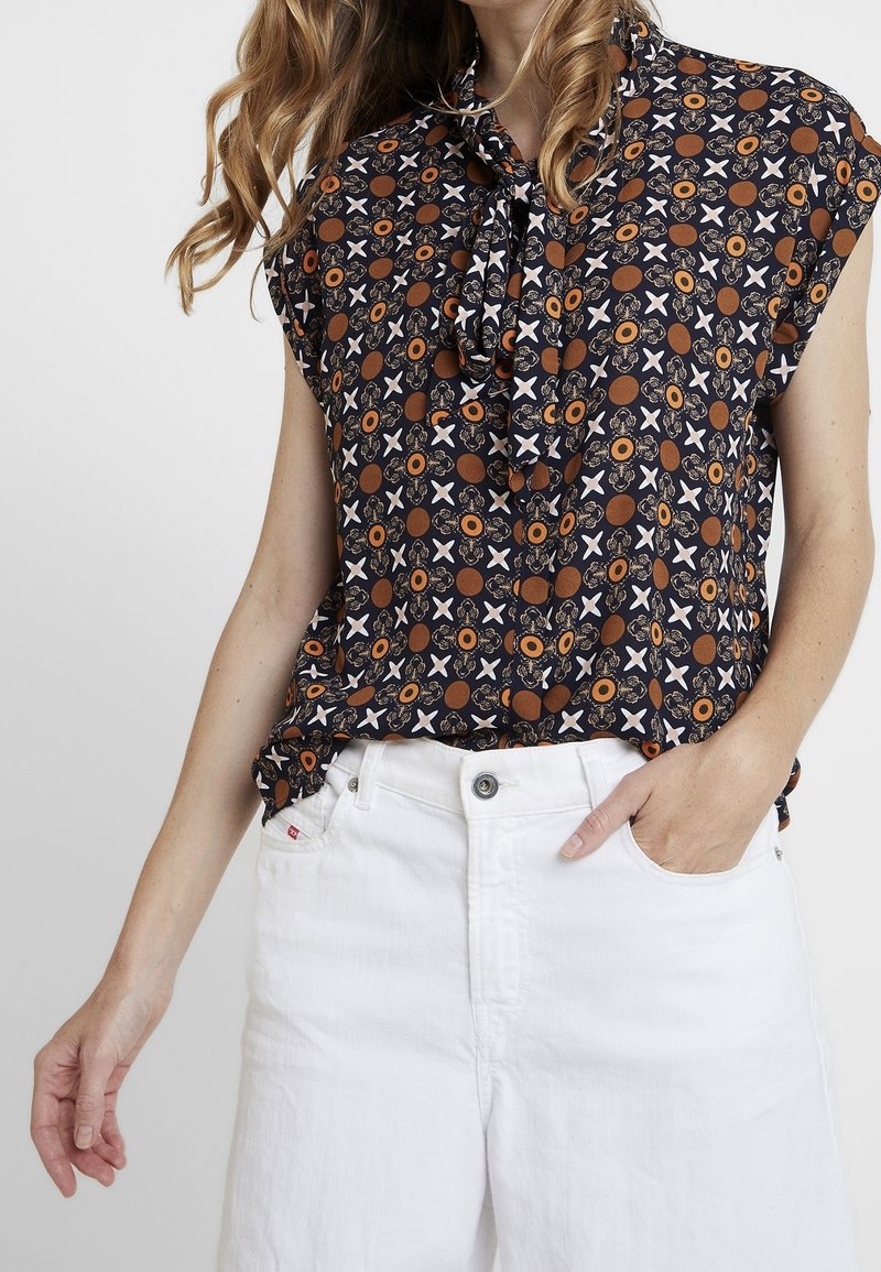 Blouse noire sans manches avec motif géométrique marron et blanc, associée à un short blanc taille haute, main dans la poche.