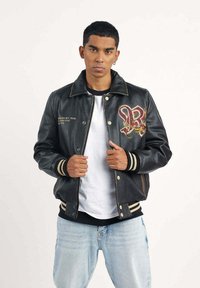 Veste bomber en cuir noir avec un design brodé "R", poignets et taille côtelés, associée à un t-shirt blanc et un jean bleu clair.