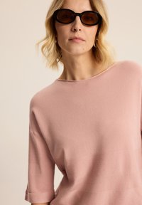 Jasnoróżowy sweter z dzianiny z rękawami o długości trzech czwartych, z teksturowanym poziomym paskiem. Model nosi ciemne owalne okulary przeciwsłoneczne.