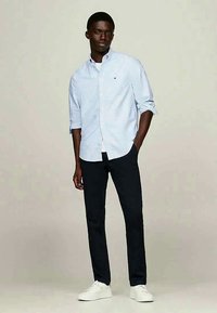 Svijetloplena karirana košulja na dugih rukava s podignutim rukavima, u kombinaciji s tamnim hlačama i bijelim tenisicama. Jednostavan dizajn i prilagođeni kroj.