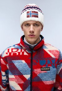 Giacca in pile a motivi rossi e blu con colletto alto e chiusura con zip; cappello lavorato a maglia bianco con patch della bandiera norvegese e accenti blu.