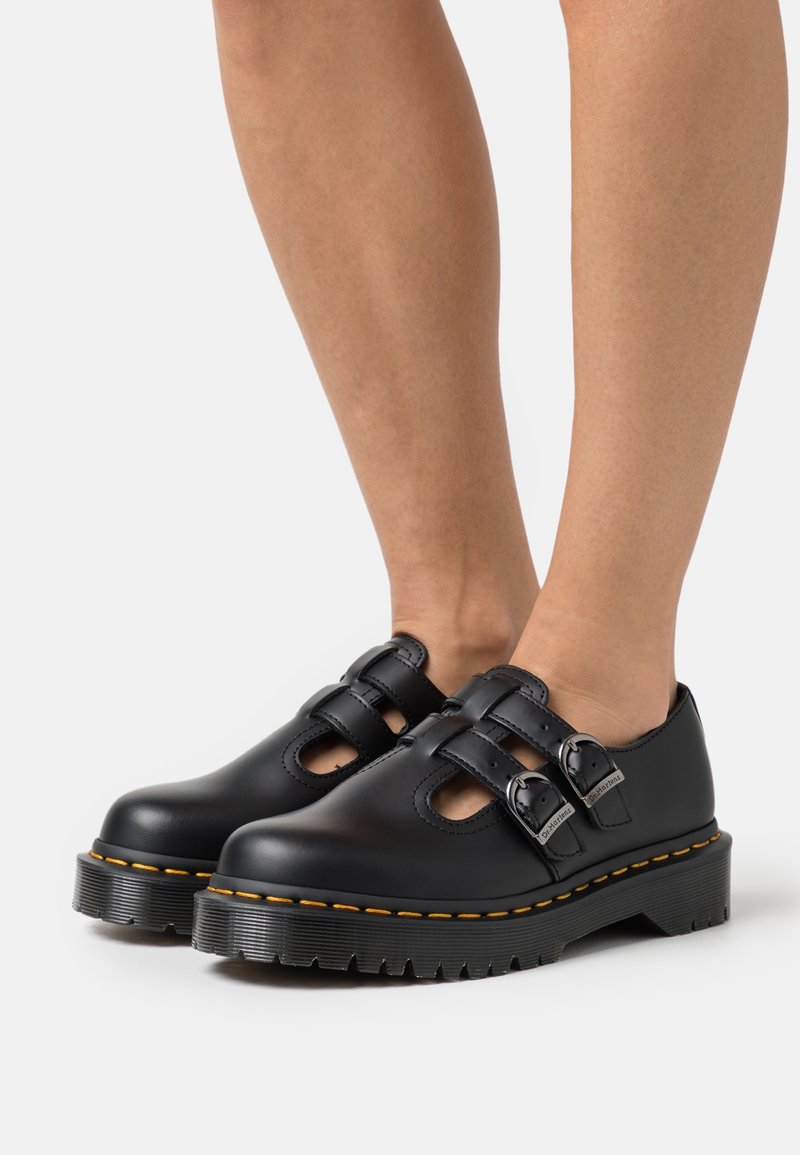 Chaussures en cuir noir avec doubles boucles et semelles épaisses crantées, portées pieds nus sur un fond blanc uni.