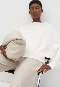 Maglione bianco oversize in maglia leggera, abbinato a pantaloni beige a vestibilità rilassata. Tiene una morbida borsa a forma di cuscino beige. Design semplice e moderno.