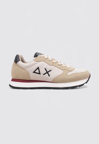 Chaussures de sport beige et crème avec des accents noirs, présentant une surface texturée, un design à lacets et un détail de semelle intermédiaire rouge.
