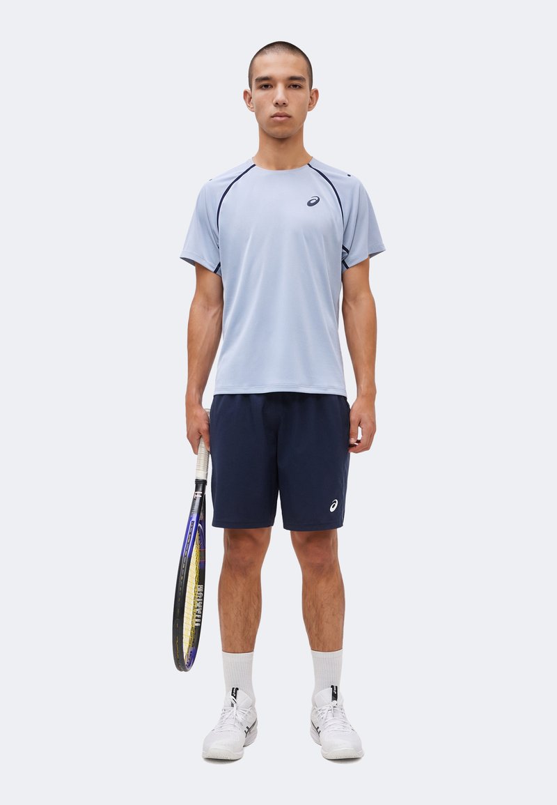 Ung mand står, holder en tennisketcher i højre hånd, iført lyseblå sports T-shirt, marineblå shorts, hvide sokker og hvide tennissko.