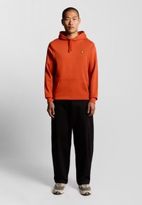 Sudadera naranja con un bolsillo canguro, capucha con cordón y pequeño logo amarillo. Combinada con pantalones negros holgados y zapatillas grises.