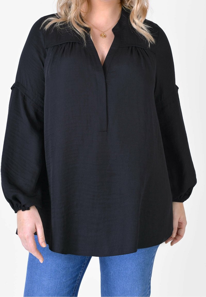 Kobieta w luźnej czarnej bluzce z długim rękawem i ściągaczami przy mankietach oraz niebieskich dżinsach, stojąca na tle gładkiego białego tła.