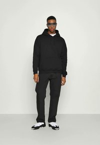 Sort hættetrøje med en forlomme, parret med sorte bukser og hvide sneakers. Model iført mørke solbriller. Minimalistisk design.