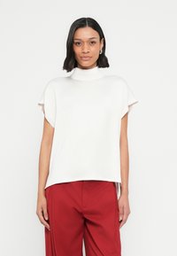 Valkoinen lyhythihainen paita, jossa korkea puolikorkea kaulus ja väljä istuvuus. Pehmeä kangas, jossa sileä pinta ja epäsymmetrinen helma. Yhdistetty punaisiin housuihin.