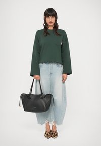 Geantă de mână din piele neagră cu suprafață texturată, două mânere și închidere cu șnur; purtată cu un hanorac verde scurt și blugi de culoare albastru deschis.