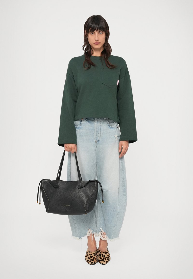 Geantă de mână din piele neagră cu suprafață texturată, două mânere și închidere cu șnur; purtată cu un hanorac verde scurt și blugi de culoare albastru deschis.