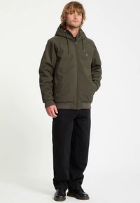 Olivově zelená bunda na zip s kapucí, elastickými manžetami a lemem, doplněná černými kalhotami s širokými nohavicemi a černými botami. Jednoduchý design a hladká textura.