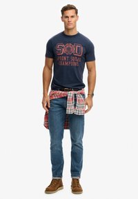Camiseta marina con estampado rojo, combinada con jeans azules y botas marrones. Una camisa de cuadros atada alrededor de la cintura añade textura y patrón.
