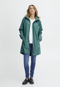 Jachetă verde impermeabilă cu glugă, accente negre, buzunare laterale, manșete elastice. Purți peste o cămașă albă și asortată cu blugi albaștri.