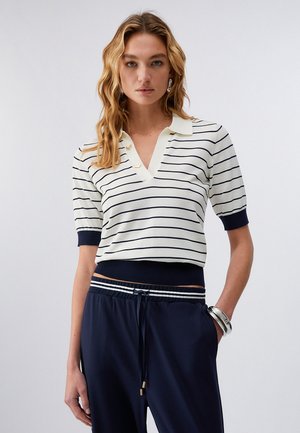 Kobieta o falowanych blond włosach ubrana w białą koszulę z krótkim rękawem, ozdobioną cienkimi czarnymi paskami i granatowymi mankietami, do tego granatowe spodnie i srebrne bransoletki.