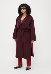 Burgundy long coat with wide lapels and a belt, featuring two buttons and side slits. Paired with matching pants and dark flat shoes.