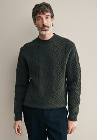 Dunkelgrüner gestrickter Pullover mit strukturiertem Zopfmuster, rundem Halsausschnitt und gerippten Bündchen. Kombiniert mit dunklen Hosen.