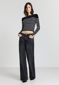 Giovane donna vestita con un top cropped a maniche lunghe a righe bianche e nere e jeans neri larghi a vita alta, in piedi con le mani nelle tasche.