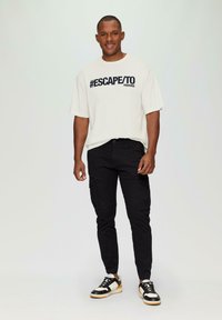 Lichtbeige t-shirt met zwarte "#ESCAPE/TO PARADISE" tekst, gecombineerd met strakke zwarte cargobroeken en zwart-witte sneakers.