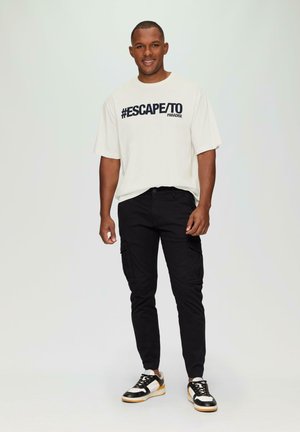 Lichtbeige t-shirt met zwarte "#ESCAPE/TO PARADISE" tekst, gecombineerd met strakke zwarte cargobroeken en zwart-witte sneakers.